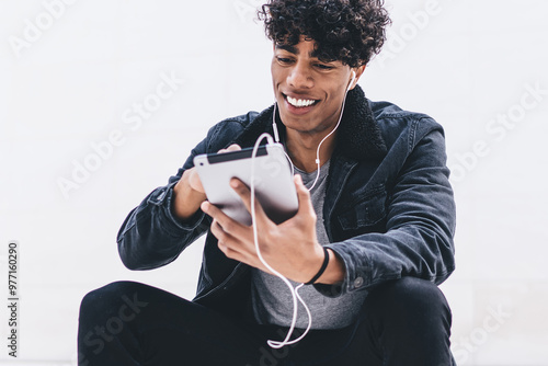 Smiling male blogger watching funny movie in headphones on internet websites via touch pad, hipster guy spending time near copy space wall for advertising while listening music playlist via app