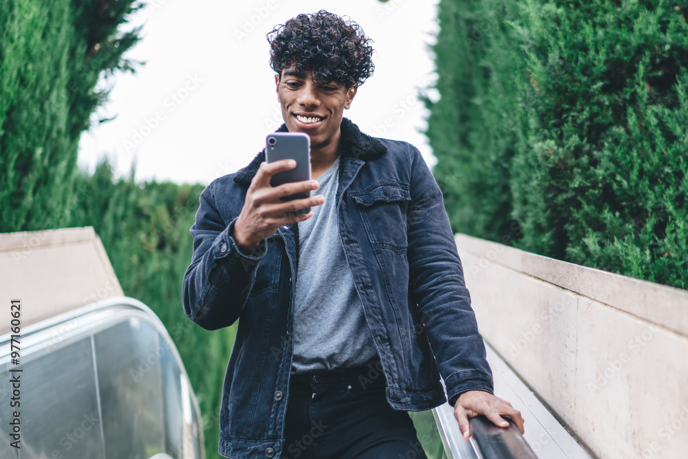 Fototapeta premium Happy hipster guy going down at city escalator and using modern cellphone gadget for online communication with followers, good looking male blogger networking while messaging on social website