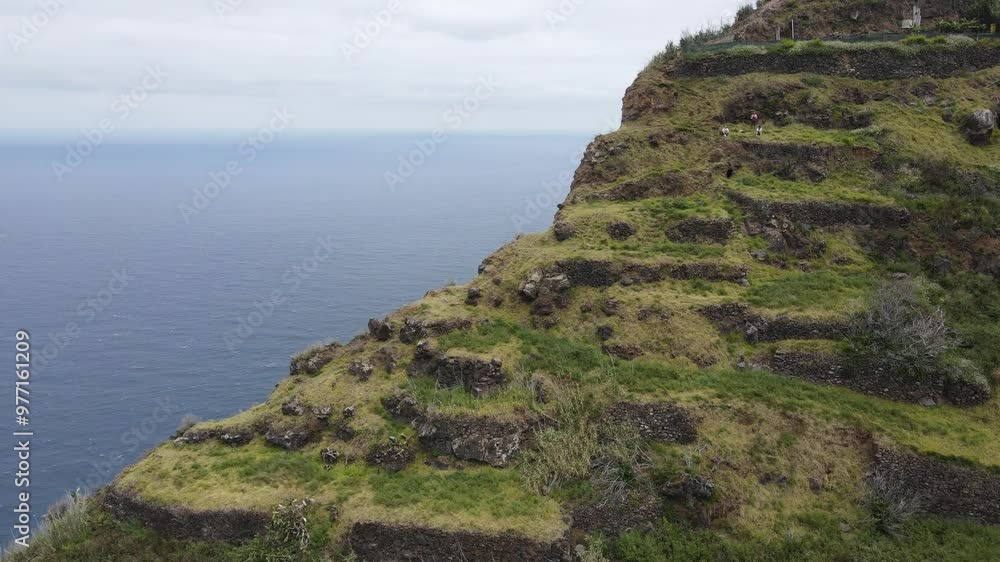 Aerial view of a steep, terraced cliff by the ocean with three hikers on the grassy terrain. The stone terraces blend with the natural landscape, highlighting rugged beauty and human presence. Aerial view of a steep, terraced cliff by the ocean with three hikers on the grassy terrain. The stone terraces blend with the natural landscape, highlighting rugged beauty and human presence.