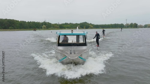 hydro foilers surfing wave behind watercraft driven by blond girl filmed with drone from front close up with trees on riverbank photo