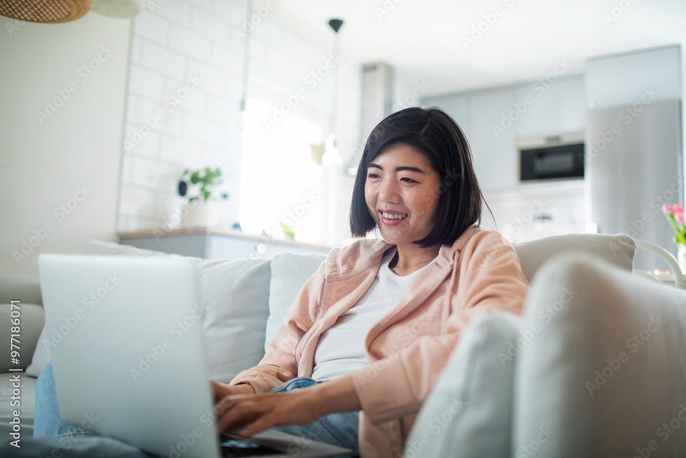 Smiling young Asian woman sitting on couch at home using laptop Smiling young Asian woman sitting on couch at home using laptop