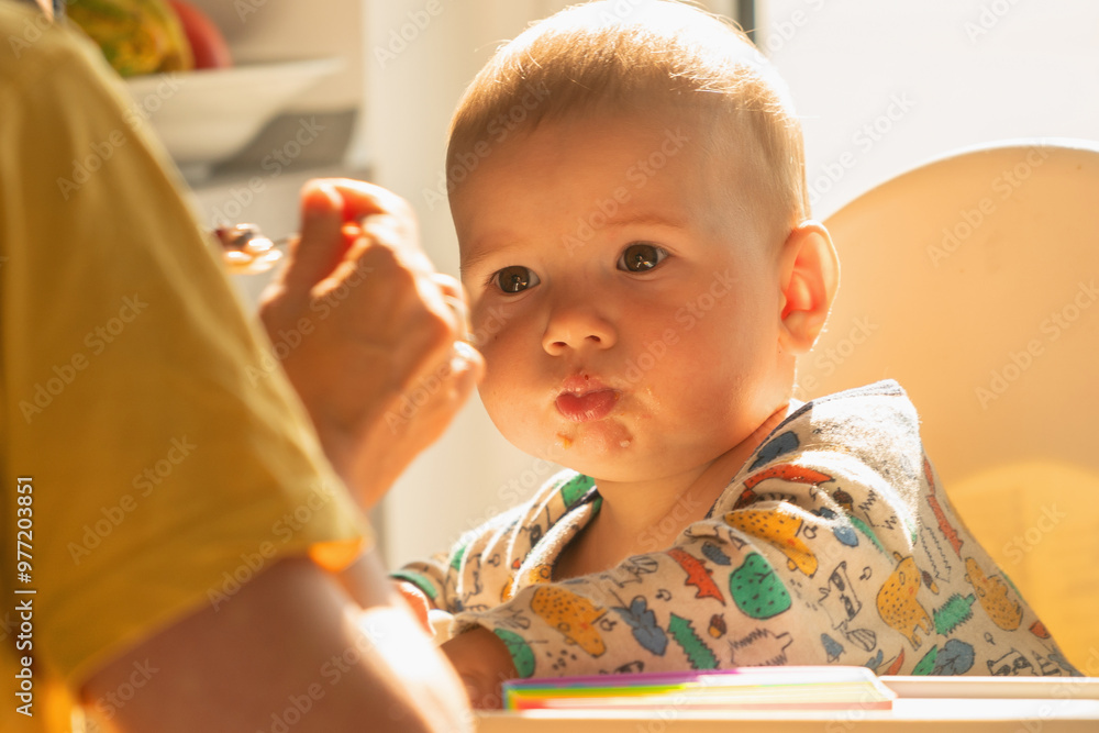 Cheerful baby being fed with porridge while sitting in white chair. Cheerful baby being fed with porridge while sitting in white chair.