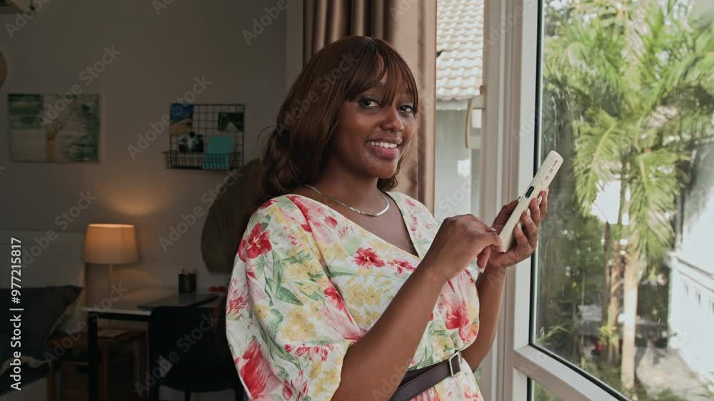 Portrait of beautiful African American girl posing by window holding mobile phone at home Portrait of beautiful African American girl posing by window holding mobile phone at home
