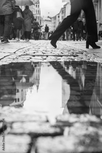 Black and white street photography capturing a rainy urban scene. A puddle reflects the historic buildings as a blurred figure steps into frame, creating dynamic movement amidst a crowded city street.