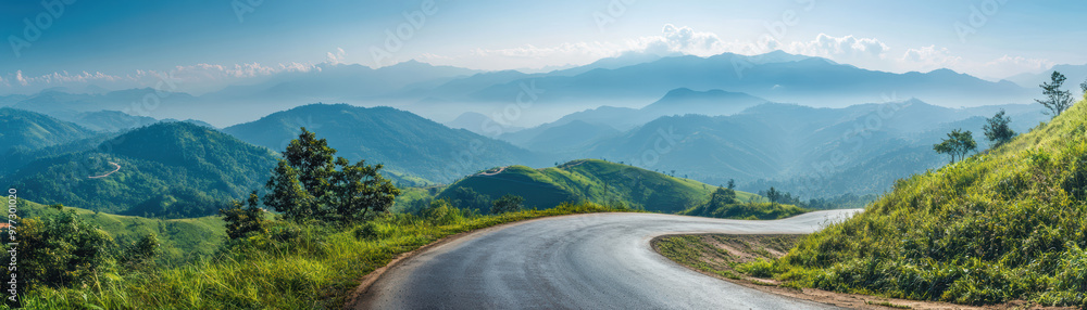 Naklejka premium Serene panoramic view of a winding road through lush green hills with misty mountains in the background under a clear blue sky. Nature at its best.