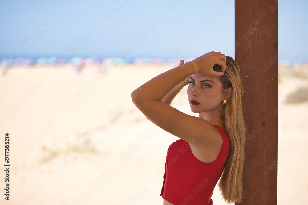 Young woman, beautiful, blue eyes, blonde, wearing red top, posing looking at camera, hands in her hair, leaning on a wooden pole with beach in background. Concept beauty, fashion, trend, femininity.