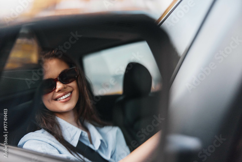 Woman in sunglasses sitting in back seat of car, hand on steering wheel, smiling