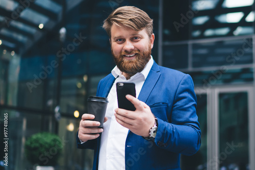 Half length portrait of cheerful male entrepreneur with takeaway cup with tasty caffeine beverage using cellphone application for checking financial news on website, concept of business people