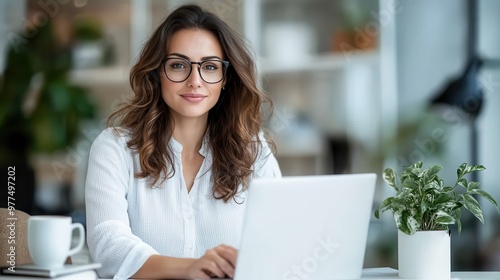 A confident woman in stylish glasses works on a laptop at a bright workspace, exuding professionalism and focus amid a cozy plant-filled environment.