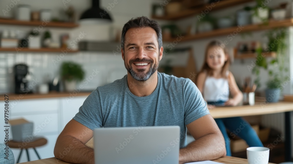 A joyful man in a gray shirt is seated at a kitchen table with a laptop in front of him, smiling as a child can be seen blurred in the background, adding a family feel. A joyful man in a gray shirt is seated at a kitchen table with a laptop in front of him, smiling as a child can be seen blurred in the background, adding a family feel.