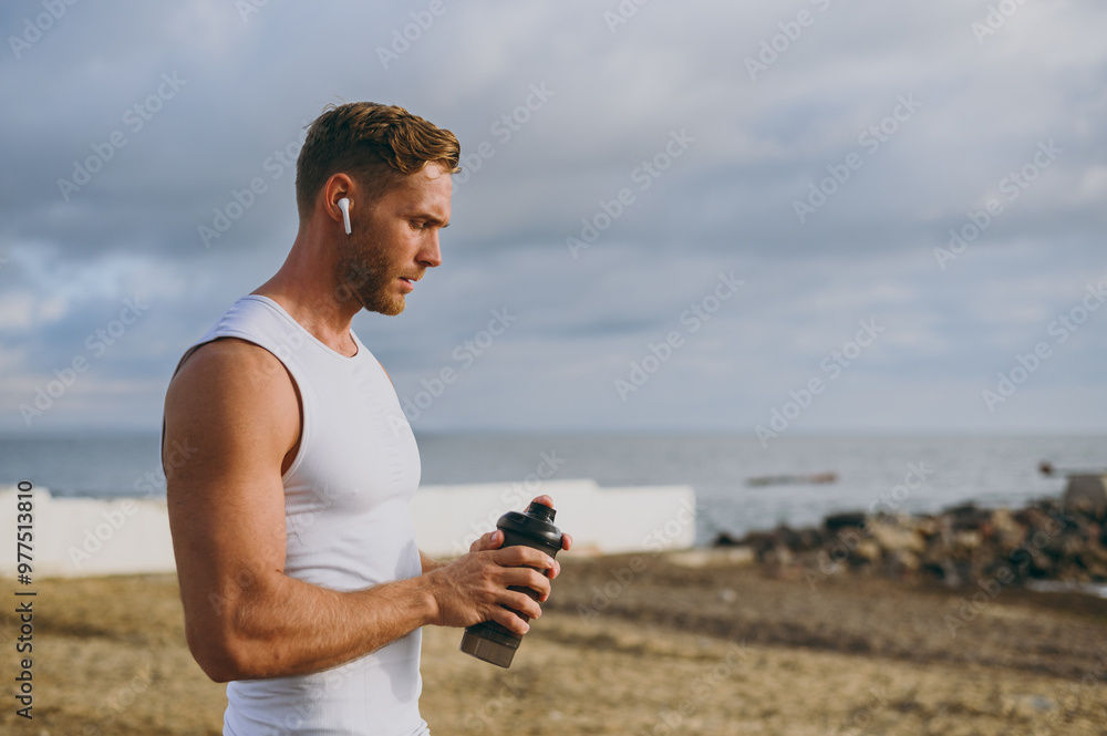 Young strong athletic toned fit sportsman man wear sports clothes earphones drink protein shake smoothie warm up training at sunrise sun dawn over sea beach outdoor on seaside in summer day morning.. Young strong athletic toned fit sportsman man wear sports clothes earphones drink protein shake smoothie warm up training at sunrise sun dawn over sea beach outdoor on seaside in summer day morning..