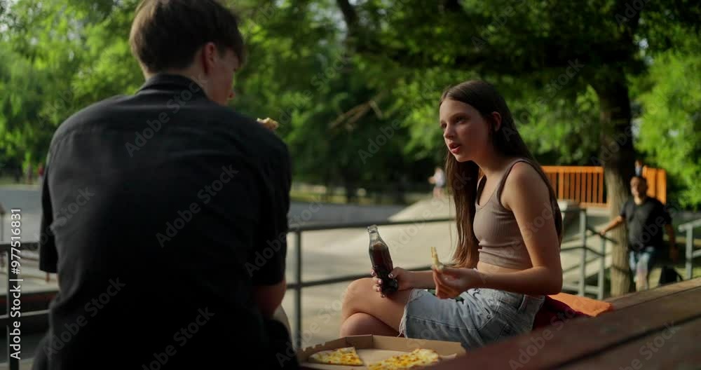 Over the shoulder a teenage girl drinks a soda while talking to her classmate guy during a meeting at a skatepark in the summer Over the shoulder a teenage girl drinks a soda while talking to her classmate guy during a meeting at a skatepark in the summer