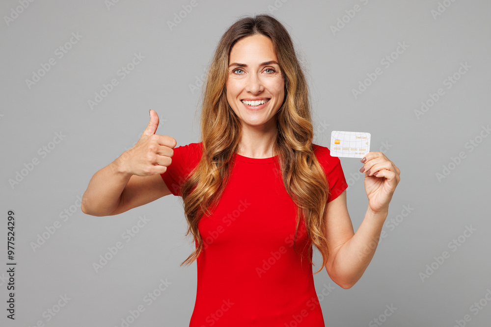 Young smiling satisfied happy woman she wearing red t-shirt casual clothes hold in hand mock up of credit bank card show thumb up isolated on plain grey background studio portrait. Lifestyle concept. Young smiling satisfied happy woman she wearing red t-shirt casual clothes hold in hand mock up of credit bank card show thumb up isolated on plain grey background studio portrait. Lifestyle concept.