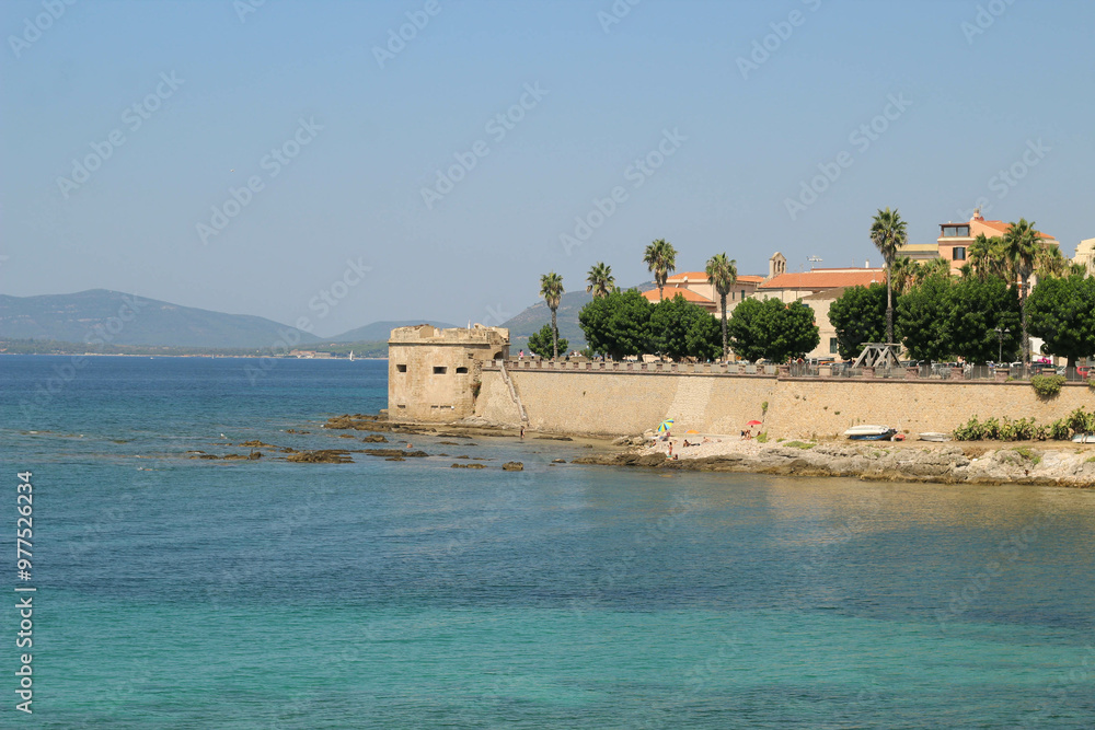 Naklejka premium A scenic view of a Mediterranean-style coastal town, featuring a historic fort or watchtower along a beach with crystal-clear turquoise waters. Palm trees, sandy shores and mountains under blue sky.