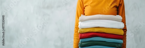 A person in a yellow shirt holding a stack of neatly folded clothes in various colors, including red, green, white, and mustard, against a light background. photo