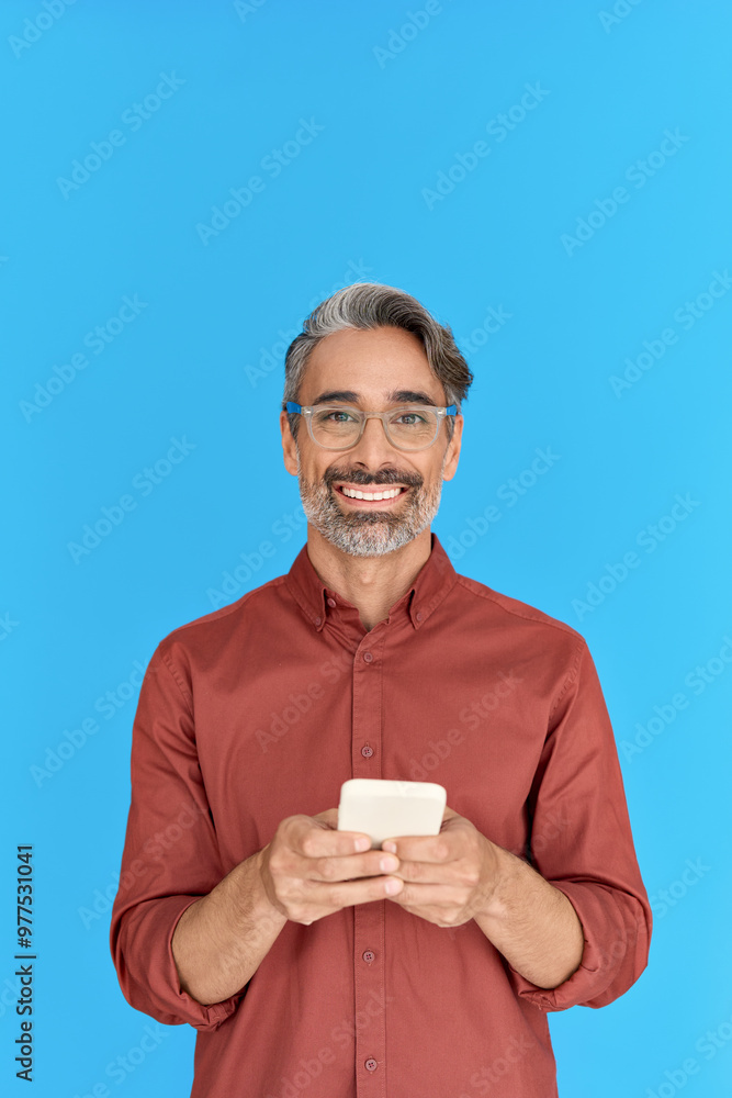 Smiling mature man using apps on cell phone standing isolated on blue background. Happy middle aged male model holding smartphone using mobile cellphone for social media apps. Vertical. Smiling mature man using apps on cell phone standing isolated on blue background. Happy middle aged male model holding smartphone using mobile cellphone for social media apps. Vertical.