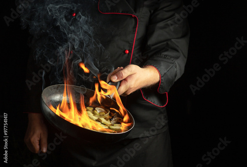 Professional chef cooking food on a frying pan with fire flames. Restaurant kitchen idea on a black background with space for advertising. Flambler or cooking vegetables with spices on natural fire. photo