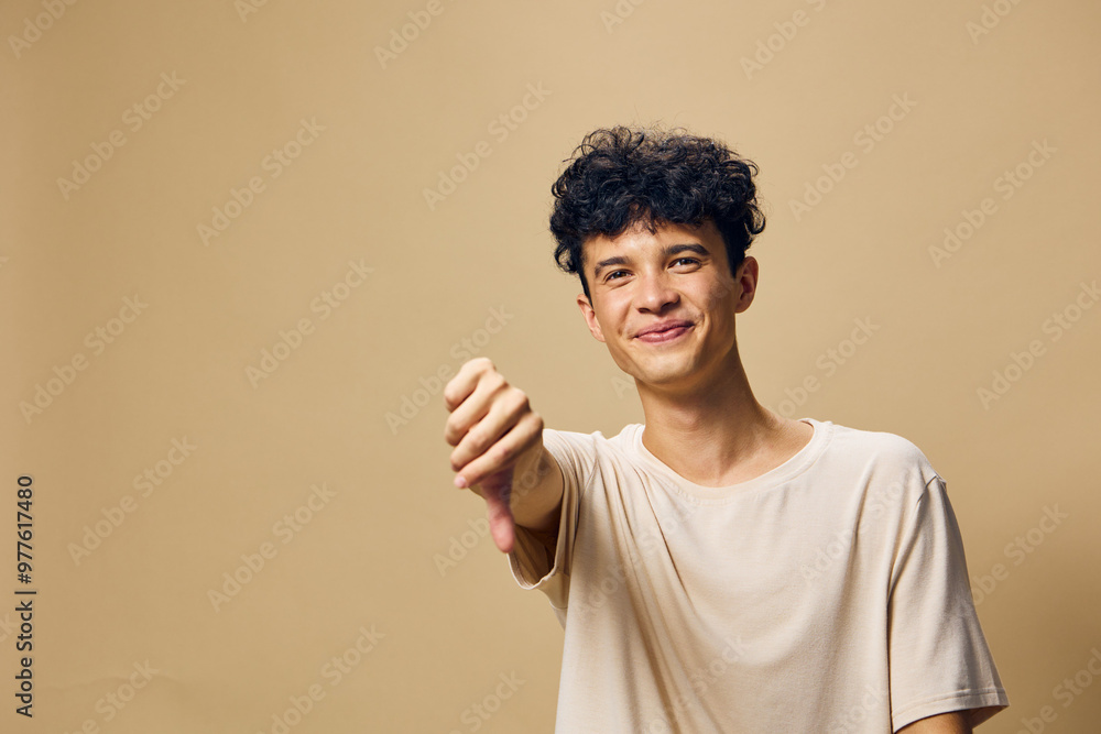 Young man expressing a thumbs down gesture in a neutral pose against a soft beige background, conveying disappointment or disapproval in a casual setting Young man expressing a thumbs down gesture in a neutral pose against a soft beige background, conveying disappointment or disapproval in a casual setting
