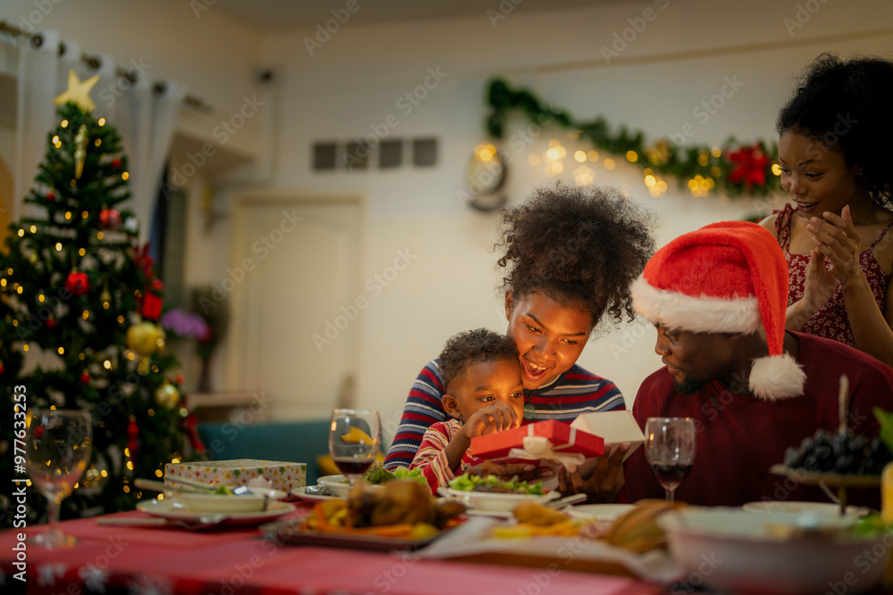A joyful family gathers around the dinner table celebrating Christmas. The father, dressed in a Santa hat, gives a gift to the child, while everyone smiles with warmth and festive decorations. A joyful family gathers around the dinner table celebrating Christmas. The father, dressed in a Santa hat, gives a gift to the child, while everyone smiles with warmth and festive decorations.