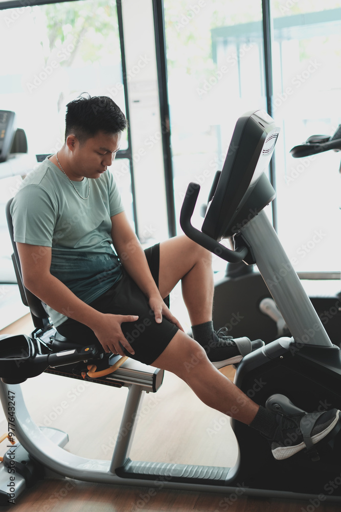Fototapeta premium This is a kind of sport injury concept; Asian male athlete sitting on spin bicycle fitness workout and lower leg cramp and backache, Sport man massaging sore muscles during training in fitness gym.