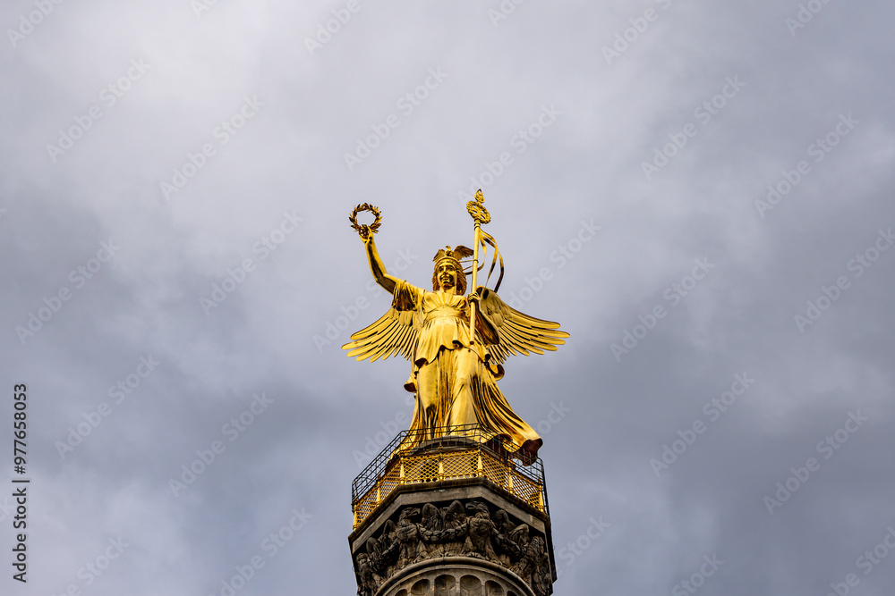 Fototapeta premium A statue of a woman holding a wreath and a bird. The statue is gold and is on top of a building. The sky is cloudy and the overall mood of the image is serene and peaceful
