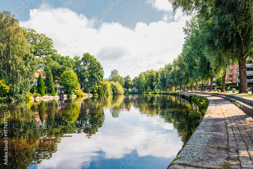 Fényképezés View of a lush riverside path in city park with tall trees and reflections in the Svartån River