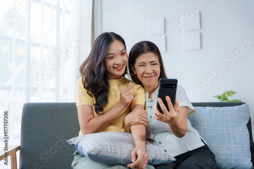 Two generations of women. An older mother and a younger daughter. Sitting on a cozy sofa in their modern home. Happily taking a selfie together