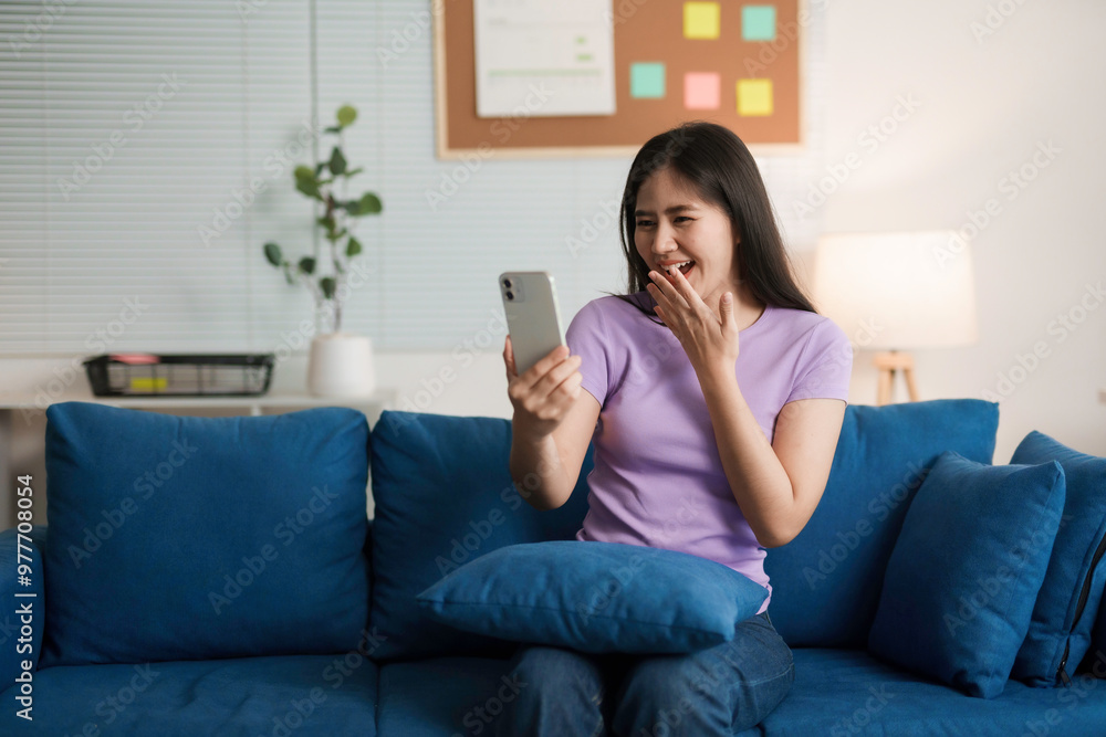Young woman smiles joyfully while video calling on her smartphone in a sunny living room, dressed in jeans and a t shirt, creating a comfortable and contemporary setting Young woman smiles joyfully while video calling on her smartphone in a sunny living room, dressed in jeans and a t shirt, creating a comfortable and contemporary setting