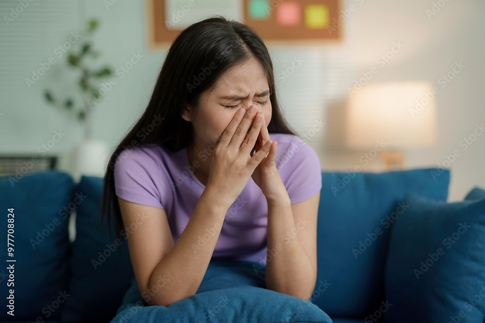 Emotional young asian woman is seen sitting on a living room sofa, with her face in her hands, visibly upset and distressed Emotional young asian woman is seen sitting on a living room sofa, with her face in her hands, visibly upset and distressed