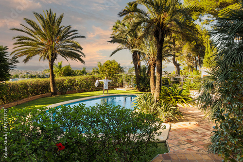 A man of retirement age in a white robe by the pool against an exotic landscape with palm trees and a pool enjoying the setting sun.
