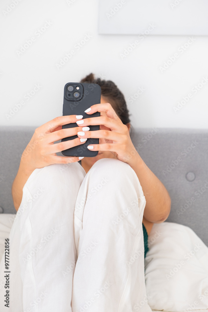 Close-up of a young teenager dressed in white linen pants sitting on her bed. She is taking a selfie for her social media and digital life with her smartphone. Teenage digital lifestyle concept Close-up of a young teenager dressed in white linen pants sitting on her bed. She is taking a selfie for her social media and digital life with her smartphone. Teenage digital lifestyle concept