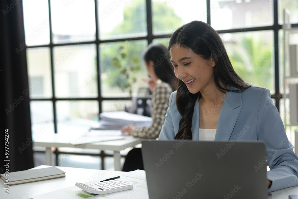 Asian businesswoman focused on her laptop work in a modern, bright office with a colleague in the background Asian businesswoman focused on her laptop work in a modern, bright office with a colleague in the background