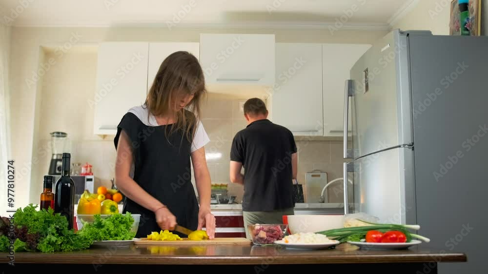 Couple preparing dinner together, wife cutting tomato with a sharp knife standing next to his husband at the kitchen counter with lying fresh leafy vegetables and bell peppers. Couple preparing dinner together, wife cutting tomato with a sharp knife standing next to his husband at the kitchen counter with lying fresh leafy vegetables and bell peppers.