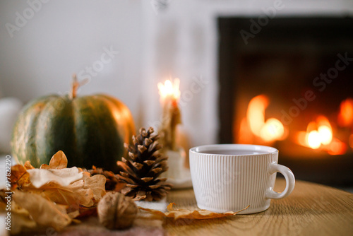 Cozy autumn. Cup of tea, pumpkin, autumn leaves on wooden table on background of warm fireplace. Fall still life in living room. Thanksgiving and Halloween.