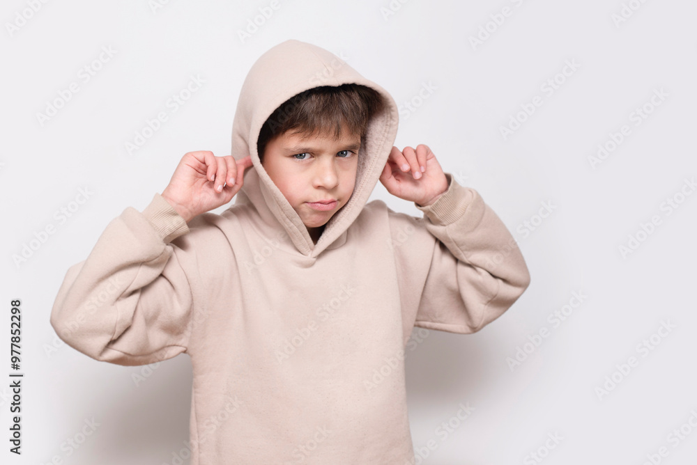 A young boy in a hoodie on a white background covers his ears and is sad, the boy is tired of constantly listening to his parents' notes, concept of education A young boy in a hoodie on a white background covers his ears and is sad, the boy is tired of constantly listening to his parents' notes, concept of education