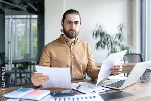 Business professional focuses on paperwork at office desk, featuring laptop, charts, and calculator. Modern workspace suggests analysis, planning, and productivity in financial or corporate setting.