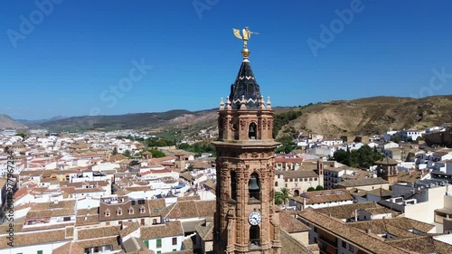 Drone fly around the belfry of an old stone church overlooking historic town of Antequera with a lot of typical andalusian white houses, narrow streets, old churches and monasteries.