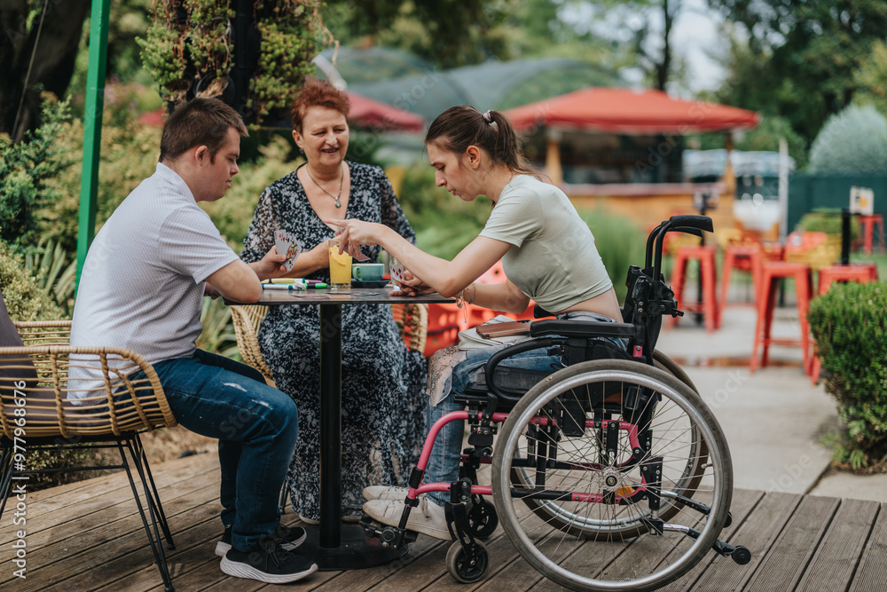 A joyful outdoor scene of a boy with Down syndrome, a girl in a wheelchair, and an older woman playing cards together, enjoying each other's company. A joyful outdoor scene of a boy with Down syndrome, a girl in a wheelchair, and an older woman playing cards together, enjoying each other's company.