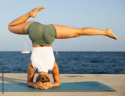 Quadro em tela Sportive young girl practicing headstand pose of yoga on blue mat on seashore