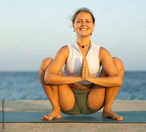 Against background of bright blue sea, slender of woman in sports clothes, hatha yoga lovers practicing oriental system of exercises. Woman performs garland pose, Malasana photo