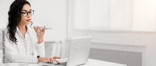 Virtual Assistant. Serious mexican businesswoman using mobile phone and working on laptop at modern office, copypace photo