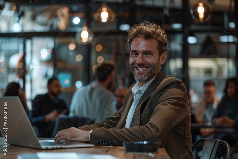 elegant man of 35 years old smiling and writing in his laptop in an office with people in the background elegant man of 35 years old smiling and writing in his laptop in an office with people in the background