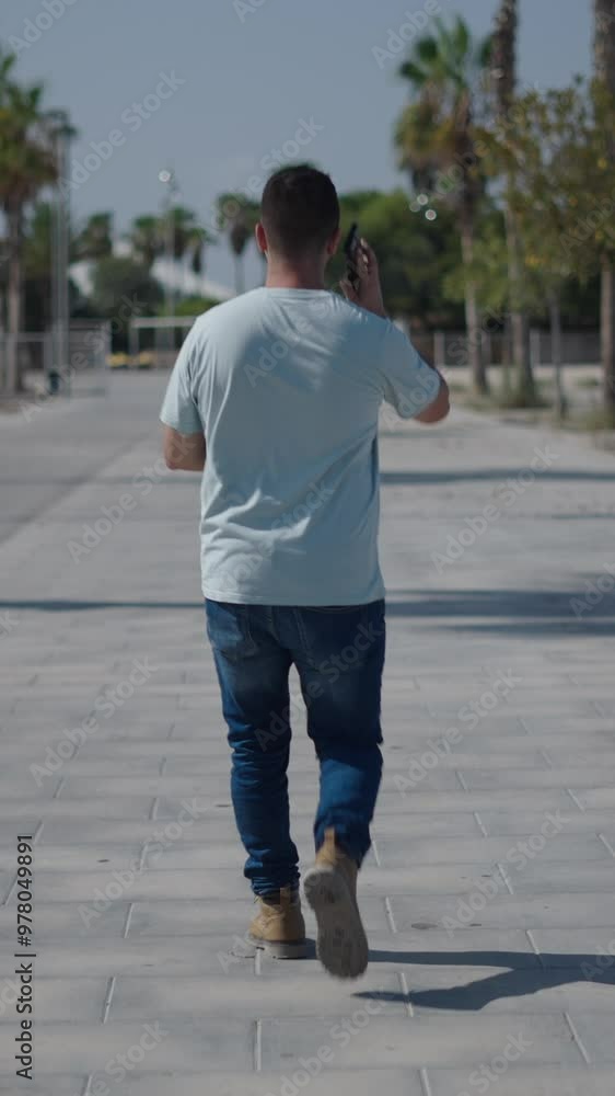 Young Caucasian boy walking with his back to the camera along the concrete walkway between the palm trees answers a call on his smartphone Young Caucasian boy walking with his back to the camera along the concrete walkway between the palm trees answers a call on his smartphone