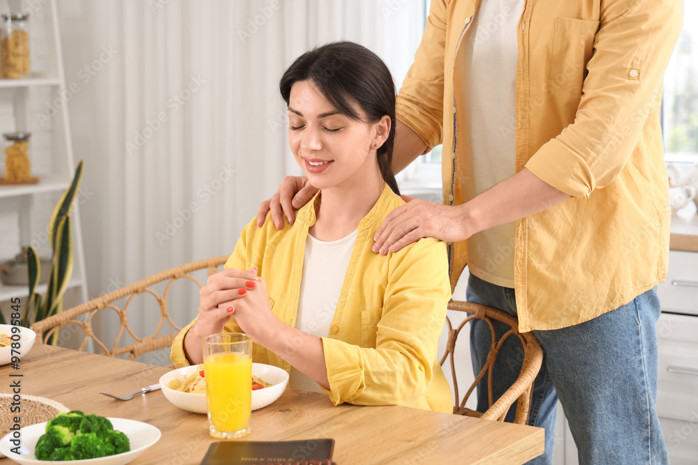 Religious young woman with her husband praying before dinner in kitchen Religious young woman with her husband praying before dinner in kitchen