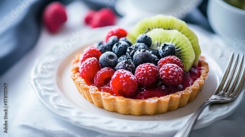 A fruit tart topped with fresh berries and kiwi slices, served on a white ceramic plate with a silver fork.