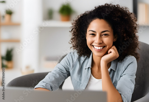 A smiling woman with curly hair sitting in a modern office, working on a laptop.