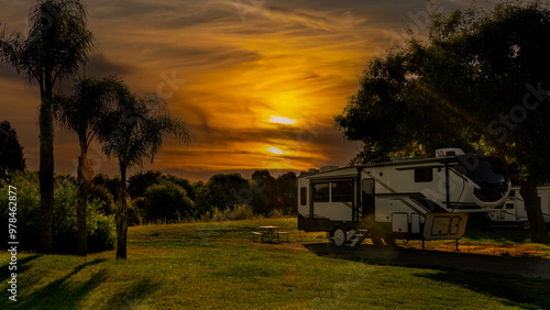 Rv motorhome fifth wheel parked at campsite under sunset sky with clouds and palm trees on grass