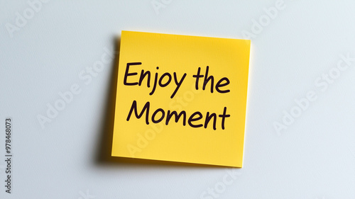 Sticky Note: A single mustard yellow sticky note with the words "Enjoy the Moment" written in simple, clear handwriting. The sticky note is on a plain white background, reminding to savor and apprecia