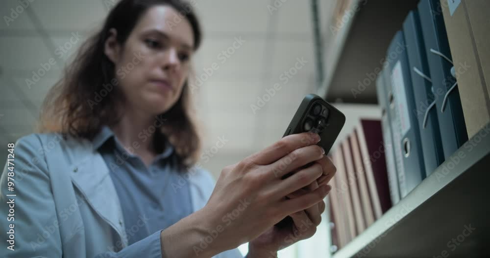 Close up of female scientist chatting, surfing the Internet on mobile phone, using AI chatbot for scientific research while standing near shelf with folders. Archaeological science lab. Low Angle. Close up of female scientist chatting, surfing the Internet on mobile phone, using AI chatbot for scientific research while standing near shelf with folders. Archaeological science lab. Low Angle.