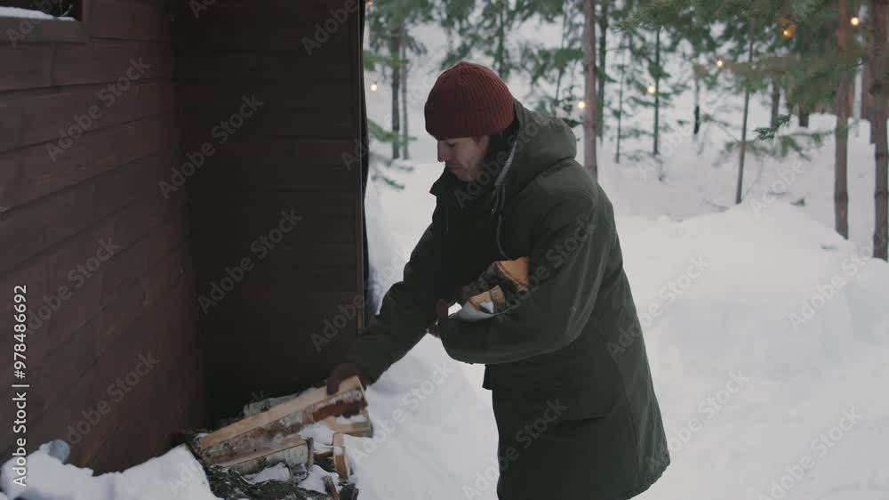 Medium full shot of young Asian man in winter clothes picking up chopped logs from wood stack outdoors by forest hut, then walking away, preparing to light fireplace Medium full shot of young Asian man in winter clothes picking up chopped logs from wood stack outdoors by forest hut, then walking away, preparing to light fireplace